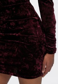 Robe en velours bordeaux avec une surface texturée, des manches longues et des fronces sur les côtés. Le tissu semble doux et brillant.