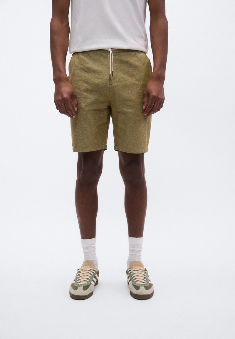 Βερμούδα από βαμβάκι σε ελαιώδες πράσινο με κορδόνι στη μέση, υφή υφάσματος, επίπεδη μπροστινή όψη και πλαϊνές τσέπες, συνδυασμένη με ανοιχτόχρωμα αθλητικά παπούτσια και κάλτσες.