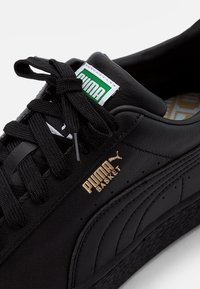 Zapatillas de cuero negro con superficie texturizada, logo dorado "PUMA BASKET", cordones negros y una etiqueta verde y blanca en la lengua.