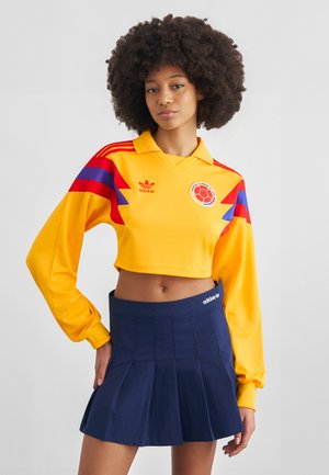 Žena s afro účesem nosí žlutý krátký fotbalový dres Kolumbie s červenými a modrými pruhy a tmavě modrou plisovanou sukní.