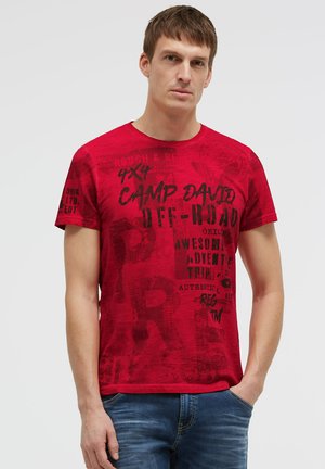 Young man with short brown hair wearing a red graphic t-shirt and blue jeans, standing against a plain light background.