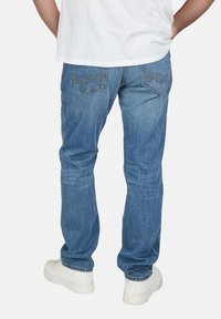 Jeans de mezclilla azul para hombre, corte regular, diseño de cinco bolsillos, pierna recta, lavado claro, detalles de costura mínimos, usados con zapatillas blancas.