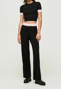 Žena v černém ribbovaném krátkém topu s krátkým rukávem a kalhotách s vysokým pasem a širokými nohavicemi s růžovými a bílými pruhy na rukávech a pásku, černé boty.