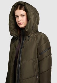Olive green puffer jacket with a large, folded hood, zip and snap closures, and a zippered sleeve pocket. Smooth texture, quilted design.