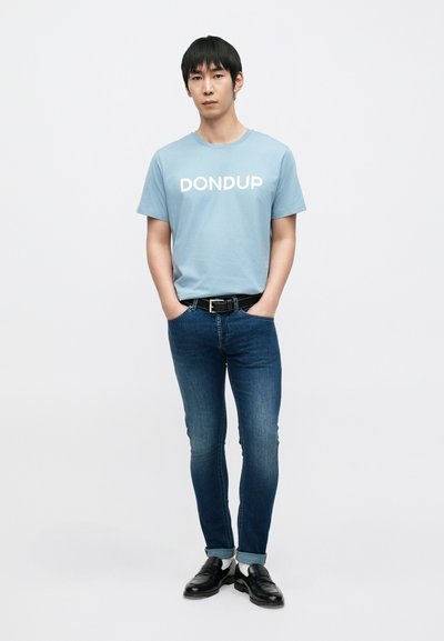 Ανοιχτό μπλε βαμβακερό T-shirt με λευκή στάμπα "DONDUP", σε συνδυασμό με σκούρα μπλε τζιν στενής γραμμής και μαύρα loafers, φορεμένα με μαύρη ζώνη.
