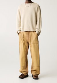 Persona che indossa un maglione crema, pantaloni cargo beige larghi con cuciture a vista e scarpe sportive marroni e nere, in piedi con le mani in tasca.