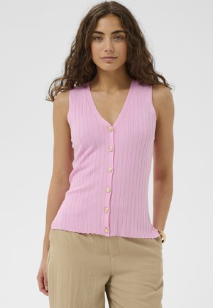 Kvinde med langt, krøllet hår iført en ærmeløs, lyserød ribbet top med knapper og beige bukser mod en ensfarvet baggrund.