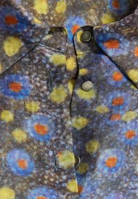 Gros plan d'un col de chemise avec boutons-pression, présentant un motif abstrait multicolore de taches bleues, jaunes et orange sur un fond gris.