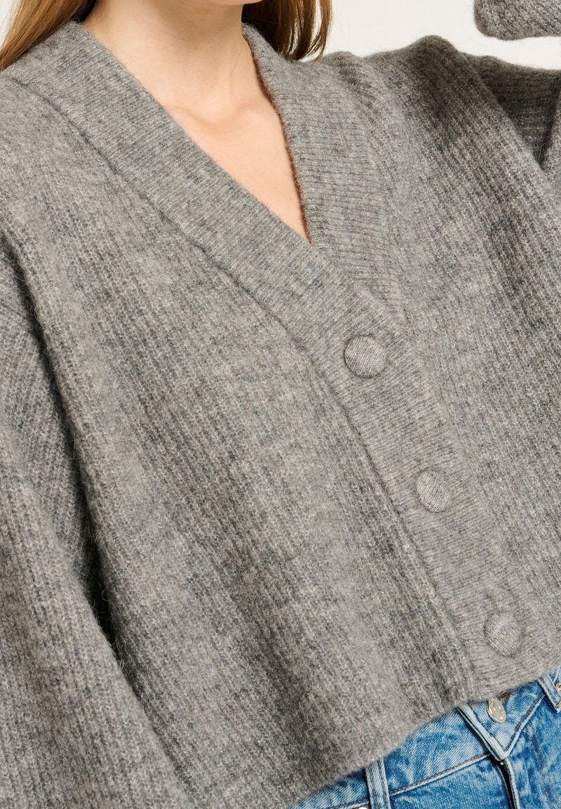 Graue Strickjacke mit V-Ausschnitt, drei Knöpfen vorne und Rippenstruktur, kombiniert mit hellblauen Jeans.