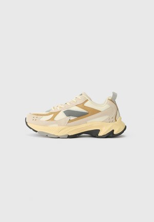 Beige en tankleurige chunky sneaker met dikke golvende zool, grijze accenten, hieltreklus en vetersluiting op een lichtgrijze achtergrond.