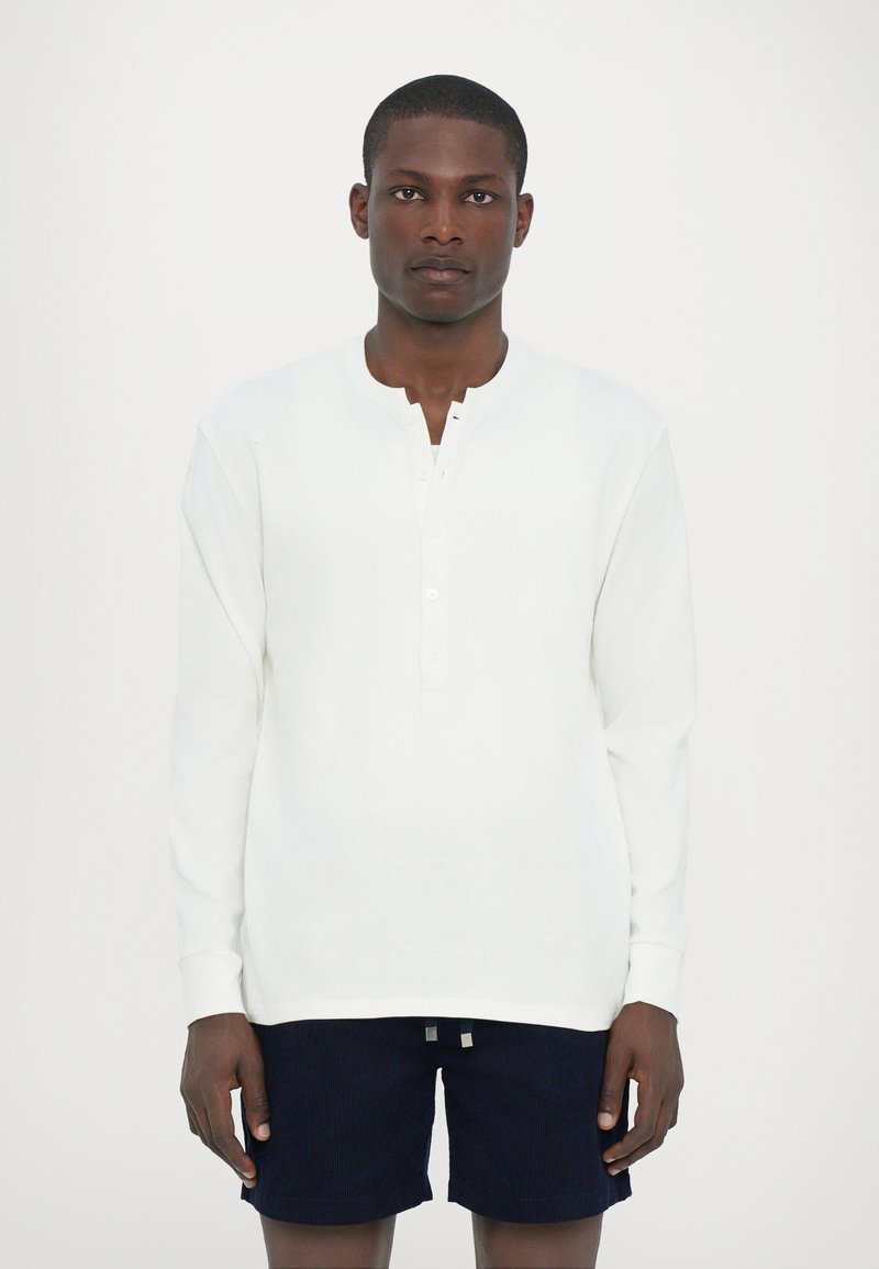 Camisa henley blanca de manga larga con cuello redondo, que presenta una placket de botones y una textura suave. Combinada con pantalones cortos de pana en color marino.