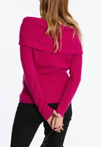 Tailliertes, schulterfreies Sweatshirt in leuchtendem Pink, aus geripptem Strickmaterial mit langen Ärmeln. Kombiniert mit schwarzen, engen Hosen.