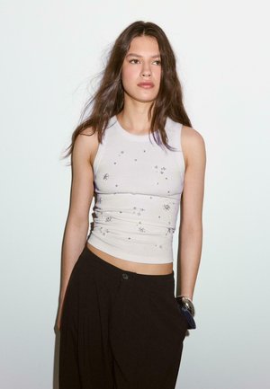 Jeune femme aux longs cheveux bruns portant un haut blanc sans manches avec de petits motifs floraux et un pantalon noir ample, debout contre un mur blanc.