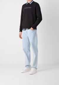 Felpa nera con il logo "Calvin Klein", sovrapposta a una camicia a righe con colletto, abbinata a jeans azzurri chiari e sneakers bianche.