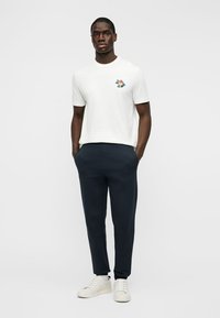 Άνδρας στέκεται με τα χέρια στις τσέπες, φοράει λευκό μπλουζάκι με μικρό πολύχρωμο γραφικό, ναυτικές παντελόνια και λευκά αθλητικά παπούτσια, με φόντο απλό.