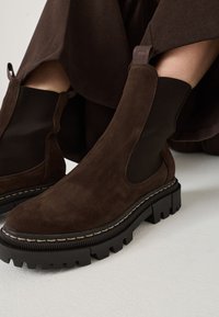 Brązowe zamszowe botki za kostkę z elastycznymi panelami bocznymi i grubymi czarnymi gumowymi podeszwami. Posiada kontrastowe przeszycie wokół podeszwy.