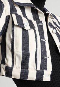 Kortärmad, kort denimjacka med blå och vita ränder, knappstängning och framfickor på bröstet med markerade sömmar.