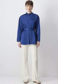 Camicia blu oversize con colletto, bottoni sul davanti e cintura abbinata; abbinata a pantaloni a gamba larga color crema e sneakers beige.