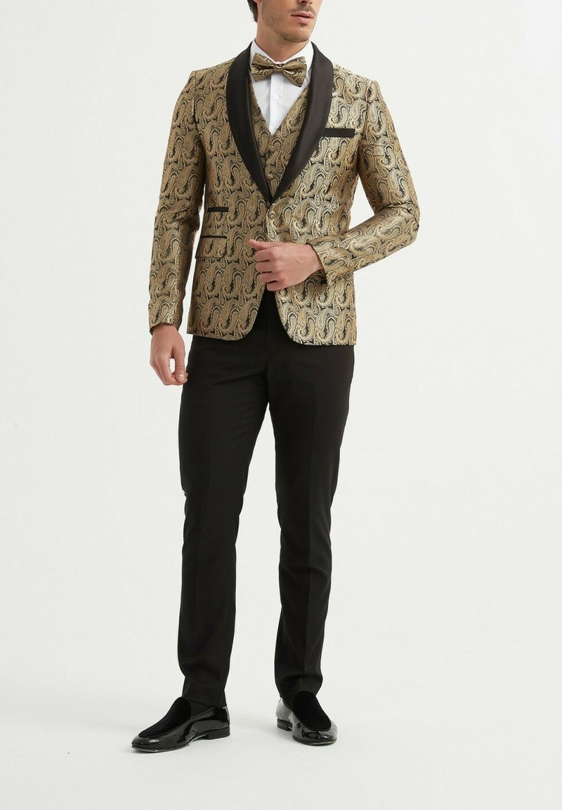 Blazer en velours doré à motif paisley, avec des revers en satin noir, associé à un pantalon noir et des mocassins brillants. Un nœud papillon complète l'ensemble.