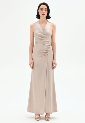 Femme aux cheveux noirs courts portant une robe beige sans manches longue jusqu'au sol avec fronces, et des talons ouverts dorés, posant devant un fond blanc.