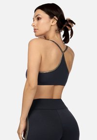 Czarny stanik sportowy z wycięciem na plecach, z złotą obwódką wzdłuż krawędzi i gładkiego materiału. Modelka jest pokazana z boku.