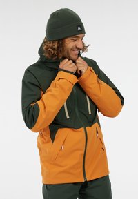 Grüne und orange wasserdichte Jacke mit einem Reißverschluss, verstellbarem Kapuze und zwei Fronttaschen. Warme Strickmütze in Dunkelgrün.