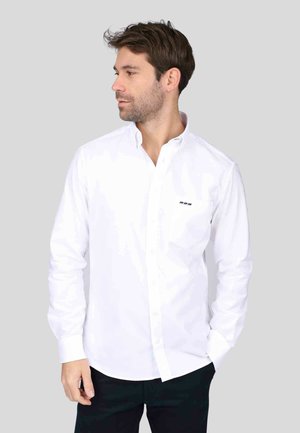 Chemise blanche à boutons, à manches longues, avec un col pointu et un petit logo noir sur la poitrine, fabriquée dans un tissu lisse.
