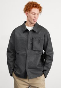 Veste chemise gris avec une finition texturée, deux poches avant et une poche poitrine zippée. Présente un col et des manches longues.