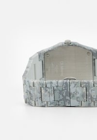 Reloj digital con una correa de patrón de camuflaje en gris y blanco, fondo de metal y esfera redonda. Presenta un diseño de enlace seguro.