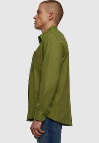 Camisa de manga larga, tipo camisa, en tela verde oliva. Tiene un bolsillo en el pecho, dobladillo recto y puños con botones.