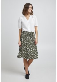 Zöld virágmintás szoknya fehér mintákkal, deréknál megkötve, fehér gombos blúzzal és fekete slip-on cipővel párosítva.