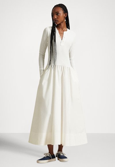 Λευκό φόρεμα μακρυμάνικο με λαιμόκοψη henley με κουμπιά, φούστα με πιέτες και τσέπες, συνδυασμένο με μπλε αθλητικά παπούτσια σε ουδέτερο φόντο.