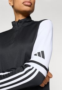 Zwart sporttop met rits en hoge hals, met witte mouwen, drie zwarte strepen op de arm en een Adidas-logo op de mouw. Glad stoftextuur.