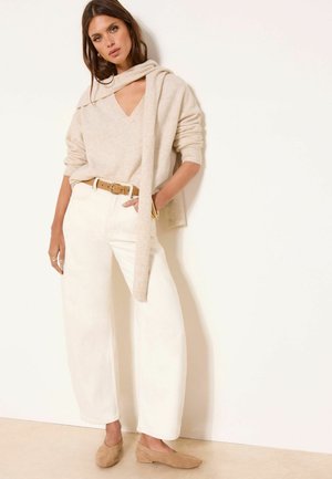 Frau lehnt an der Wand, trägt einen beigen Pullover mit passendem Schal, weiße weite Hosen, einen braunen Gürtel und beigen Ballerinas.