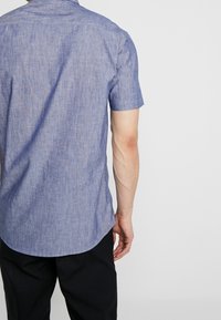 Korte mouwen, lichtblauw overhemd van gestructureerde stof, met een afgeronde zoom en een eenvoudige kraag. Achteraanzicht toont geen opvallende patronen of accenten.