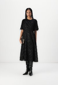 Robe noire à mi-longueur avec un tissu texturé, manches bouffantes courtes, et un motif en chevrons. Associée à des bottes noires jusqu'aux genoux et un sac à main.