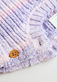 Tissu en peluche texturé violet, rose et blanc avec des stries horizontales, comportant un bouton en bois étiqueté "SUIVANT" et un élément de design coordonné.