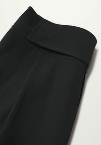 Manche en tissu noir avec un large poignet présentant un détail de couture et une fermeture à rabat rectangulaire sur fond blanc.