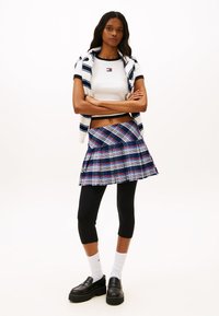 Λευκό κοντό τοπ με μαύρο τελείωμα, μπλε ριγέ φούτερ με κουκούλα, πλισέ καρό φούστα σε ναυτικό μπλε, κόκκινο, πράσινο, μαύρα κολάν και χοντρά μαύρα παπούτσια.