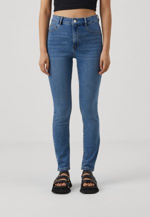 Kvinde iført højtaljede blå skinny jeans og sorte flade sandaler, stående mod en ensfarvet lys baggrund.