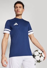 Navy blue athletic t-shirt with white striped sleeves and an Adidas logo on the chest. Model holding a patterned soccer ball.
