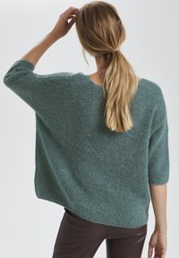 Loose-fit teal sweater with three-quarter sleeves, chunky knit texture, and a rounded neckline, paired with dark fitted pants.