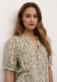 Top beige chiaro con un motivo marrone a spirale, maniche corte e scollatura a V. Realizzato in un morbido tessuto testurizzato.