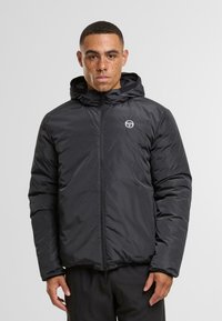 Chaqueta aislante negra con capucha, que cuenta con un cierre frontal y bolsillos laterales. Logotipo sutil en el pecho. Textura suave y diseño acolchado.