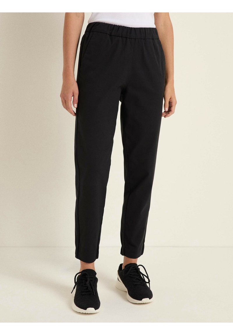 Personne portant un pantalon noir fuselé avec une taille élastique et des baskets noires, debout sur un sol clair contre un mur uni.