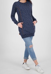 Túnica de manga larga en color navy con estampado de pájaros, que presenta bolsillos laterales y ribetes en marrón contrastante. Combinada con jeans de mezclilla claros y desgastados y zapatillas blancas.