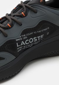 Chaussure de sport noire avec une tige en mesh texturé, des accents en caoutchouc gris, une semelle noire et des détails orange. Présente le logo de la marque et des coordonnées imprimées.