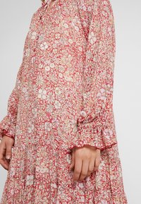 Röd blommig klänning med en lagerdesign, långa ärmar med volangdetaljer och lätt tyg. Mönstren inkluderar olika blommor.