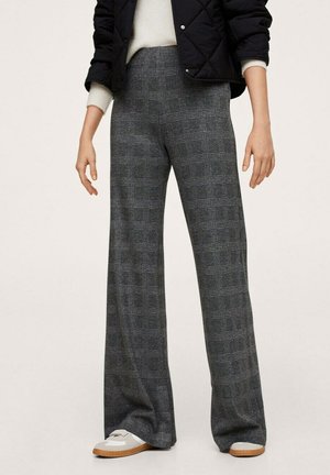 Femme portant un pantalon ample à taille haute gris à carreaux, des baskets blanches et beiges, un pull blanc et une veste matelassée noire.