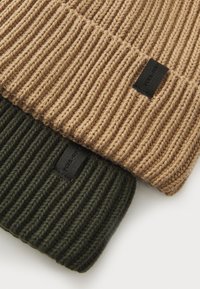 Two ribbed knit beanies, one in light brown and the other in dark green, featuring a small black label on each. The texture appears thick and warm.